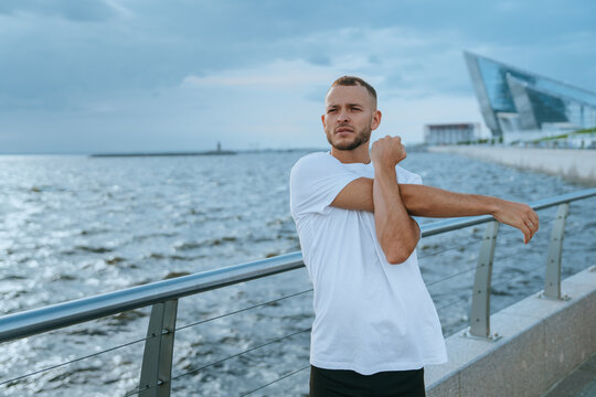 A man in a white t-shirt stretches by a waterfront, under a cloudy sky, preparing for exercise.