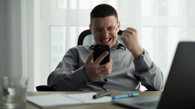 A happy man reacts to good news on his smartphone, celebrating a victory.
A surge of positive emotions emanates from a young businessman sitting at a desk in a bright room.