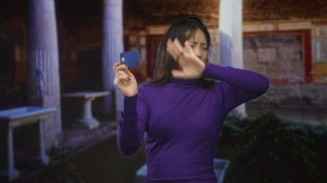 Woman holds creditcard in hand, waves other hand across face and frowns in building; financial concern annoyance.