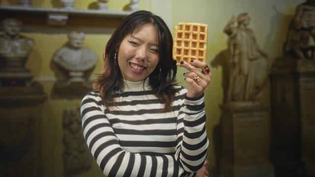 Young chinese woman holding a waffle up near her face, smiling with eyes closed, striped sweater and nose ring, statues visible in building; playful leisure.