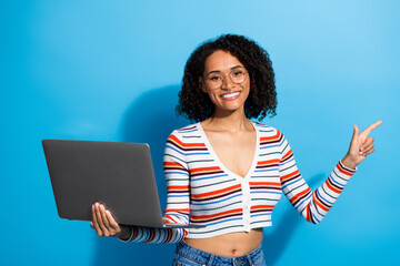 Young chic woman with glasses holds laptop and points on blue background