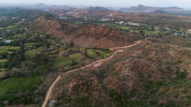 Aerial view of the Aravalli hills near Udaipur, Rajasthan at sunset, showing winding roads, patchwork agricultural fields, native trees, and scattered village houses across rugged terrain.