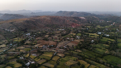 Obraz premium Aerial view of the Aravalli hills near Udaipur, Rajasthan at sunset, showing winding roads, patchwork agricultural fields, native trees, and scattered village houses across rugged terrain.