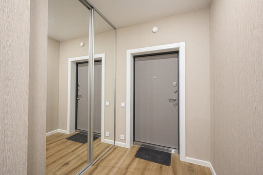 A modern hallway with beige textured walls, light wood laminate floor, metallic entrance door, sliding mirror closet, dark mats, white door frames, minimalist design.