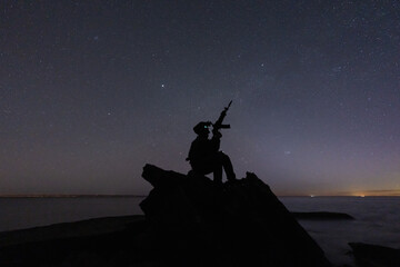 Silhouette of a lone tactical operator with night vision sitting on a rock near the ocean, holding a rifle under a vast starry sky. © Dmitri