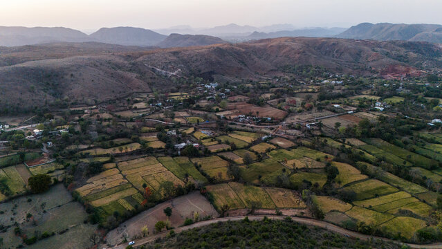 Aerial view of the Aravalli hills near Udaipur, Rajasthan at sunset, showing winding roads, patchwork agricultural fields, native trees, and scattered village houses across rugged terrain.