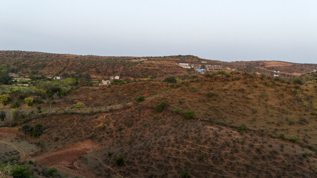 Aerial view of the Aravalli hills near Udaipur, Rajasthan at sunset, showing winding roads, patchwork agricultural fields, native trees, and scattered village houses across rugged terrain.