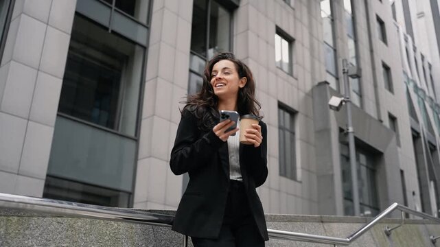 Beautiful Caucasian girl with long dark hair standing outdoor, smiling, scrolling her smartphone, drinking hot drink from eco paper cup to go.