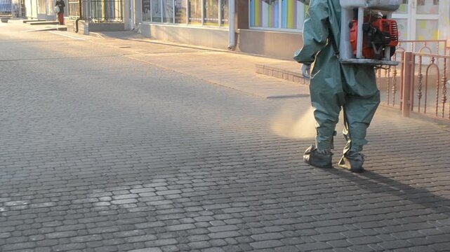 Man in airtight suit sprays disinfecting pavement on street in city. Sanitation workers clean crosswalk. Sanitary measures. Coronavirus pandemic Knapsack sprayer
