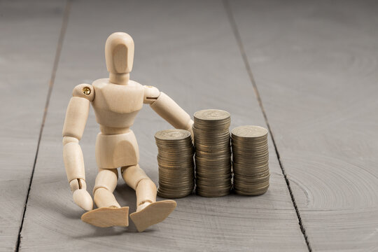 Wooden mannequin seated beside three stacks of coins, wealth and savings concept