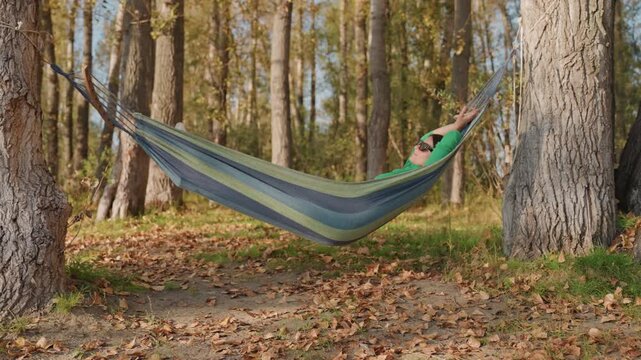 Woman relaxing in hammock among birch trees, camping nap in quiet forest green sweater peeks from woven fabric, gentle sway above leafstrewn ground, warm sunlight filtering through trunks, peaceful