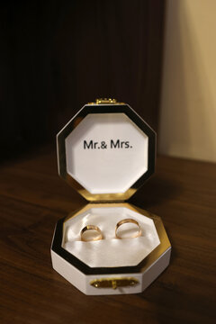 Golden wedding bands placed in an octagonal ring box with &ldquo;Mr. & Mrs.&rdquo; lettering, set on a wooden surface in a cozy, warmly lit room.