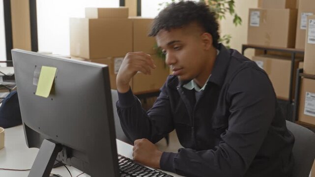 Man at computer typing with hand to head among stacked parcels and boxes in office building; fatigue duty.
