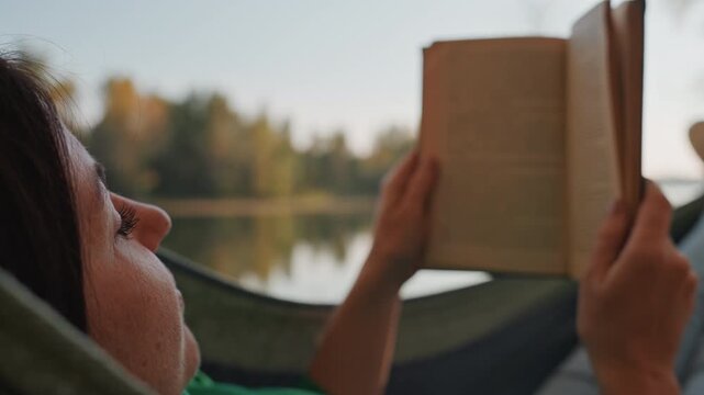 Closeup hammock reading caucasian woman camping, solo traveler focusing on text, morning light over lake, subtle breeze, fingertips turning pages, soft sweater texture, concentrated mood, quiet escape