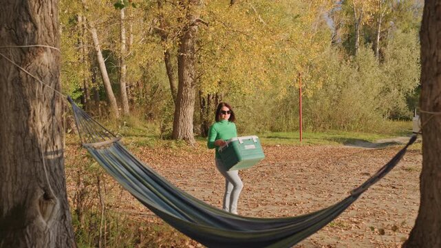 Solo black woman adjusts hammock at campsite wearing green sweater and sunglasses, walks across leaf covered clearing, ties hammock between trees near lake, parked suv in background, warm golden light