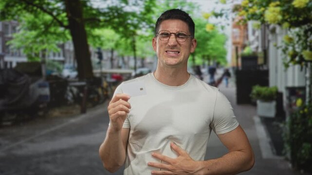 Man holding creditcard and clutching stomach on a busy street sidewalk, grimacing while inspecting the card; card trouble pain.