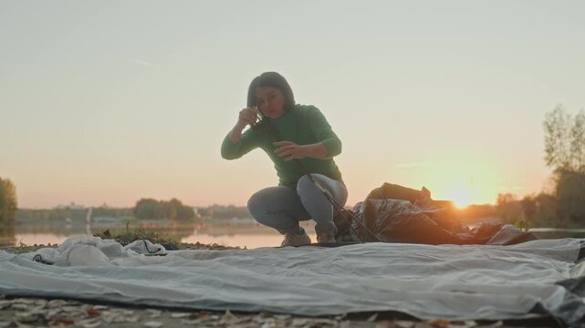 Woman setting up tent on lakeshore, crouching on groundsheet and laying poles while checking knots and gear, warm golden sunset over calm water, treelined horizon, peaceful reflective mood, preparing