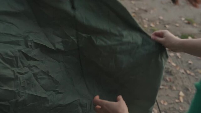 White woman securing tent base on sandy shore, fastening clips and pegs, tucking edges, pressing stake into soil, denim jeans and sneakers visible, autumn leaves underfoot, closeup motion detail,