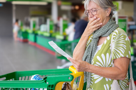 Worried senior woman standing in a supermarket checkout area, holding and reviewing her shopping receipt beside a grocery cart, expressing concern about household expenses and rising food prices