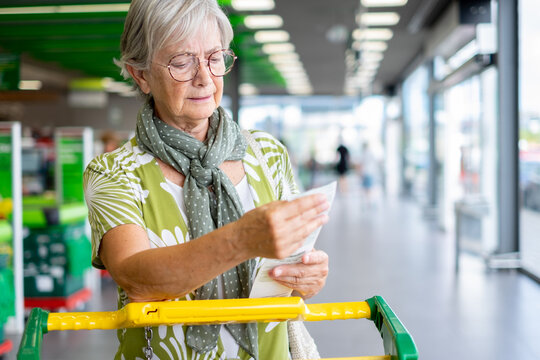 Senior woman standing in a supermarket checkout area, holding and reviewing her shopping receipt beside a grocery cart, expressing concern about household expenses and rising food prices
