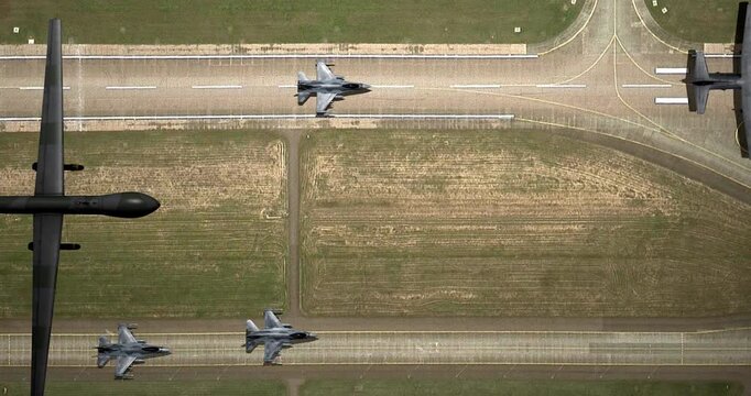 Drone flies over a runway where fighter jets are moving.
An overhead view of a drone carrying bombs flying past and several fighter jets preparing for takeoff, casting shadows on the ground.
