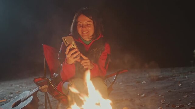 Smiling caucasian camper by campfire snack. Unwrapping chocolate bar, warm laugh relaxed posture, red chair, guitar leaning nearby, sparks rising, night sky overhead, content cozy vibe