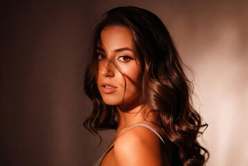 Naklejka premium A young woman with long, wavy hair is gazing slightly to the side. She’s in a studio setting with warm lighting and a textured wall background.