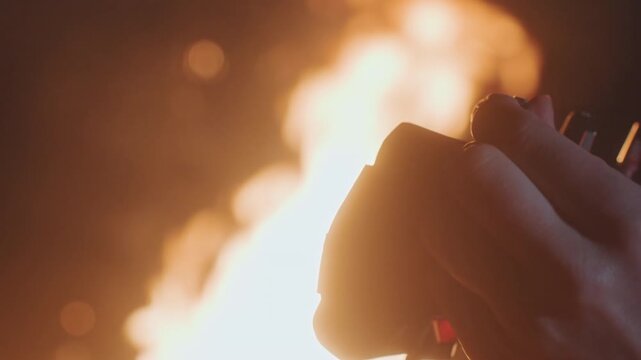 Closeup hands tuning guitar by campfire, warm orange glow and sparks, headstock and strings in sharp focus, fingers adjusting pegs, evening outdoor mood, textured jacket sleeve and ember bokeh