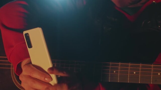 Closeup white woman holding phone near guitar and campfire, red jacket, warm orange glow, checking strings and adjusting tuning, introspective expression, cozy nighttime camping mood
