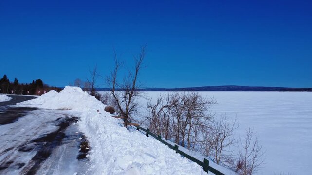 Drone footage of Lake Matapedia, Quebec. A quiet winter scene featuring deep snowbanks, bare trees, and a vast frozen lake under a crisp blue sky. Capturing a cold, remote Canadian atmosphere. 