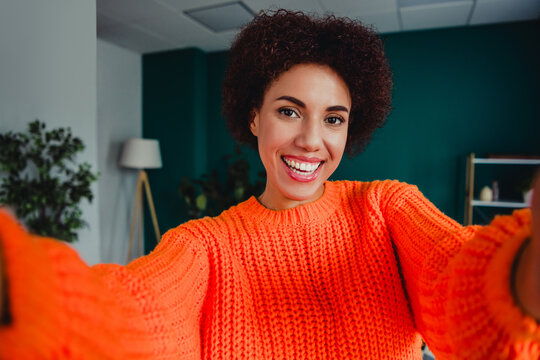 Happy young woman taking a selfie at home wearing a bright orange sweater and smiling in a cozy home office portrait
