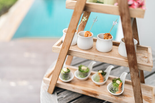 Appetizer finger food snack catering party outdoor table wooden stand ceramic cup swimming pool background, elegant bite size food display rustic serving stand beside pool for summer party