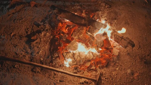 Closeup embers on sandy ground at campsite stick pokes glowing coals, smoldering cinders and smoke, sparks drifting, camper tending fire for warmth and cooking, focused maintenance, intimate texture