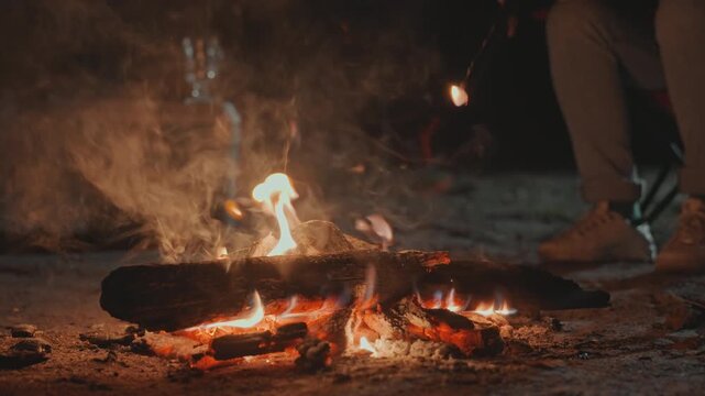 Seated camper stoking fire near kettle. Visible legs and boots beside burning logs, steam rising from metal kettle as stick pokes embers. Warm glow casts long shadows across campsite, subtle smoke