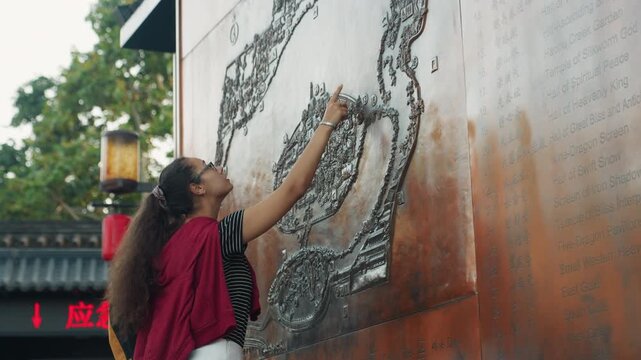 Young woman touching metal relief wall, outdoor temple courtyard, red shawl and striped shirt, long curly hair and glasses, tracing raised bronze pattern with fingertip, curious expression, warm