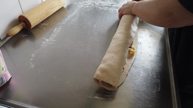 Hands roll a filled dough log on a stainless steel kitchen counter, illustrating the baking process, homemade pastry preparation and hands-on cooking in a home kitchen setting.