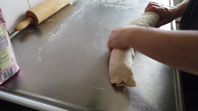 Hands roll a filled dough log on a stainless steel kitchen counter, illustrating the baking process, homemade pastry preparation and hands-on cooking in a home kitchen setting.