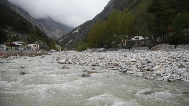 Scenic mountain river flowing through a rocky valley surrounded by pine trees & mist-covered hills. Peaceful natural landscape with flowing water, stones & small hillside settlement in the background.