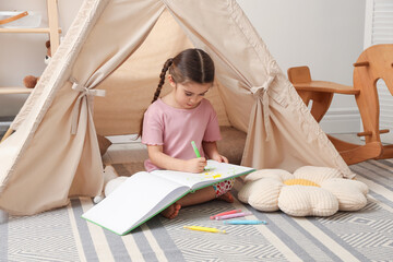 Little girl drawing in sketchbook near toy wigwam at home © New Africa