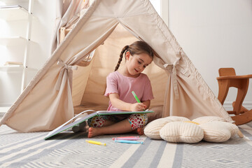 Little girl drawing in sketchbook near toy wigwam at home © New Africa