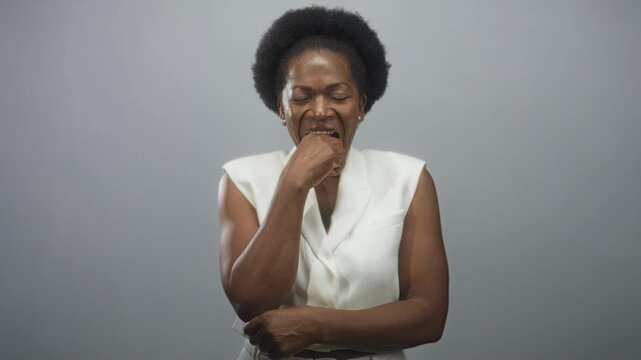 Woman biting her fist with eyes closed in studio, wearing white sleeveless waistcoat and belt, expression of tension and nervousness; anxiety coping resilience.