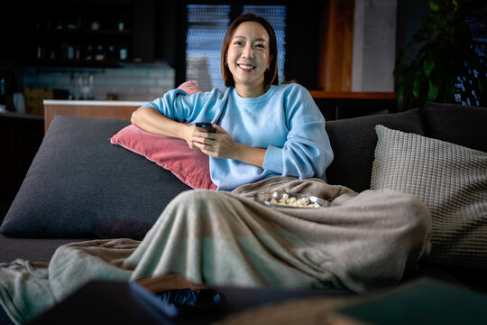 East Asian woman relaxing on a sofa at night, holding a TV remote and smiling while watching something entertaining. Cozy evening scene with popcorn, blanket, and warm home atmosphere
