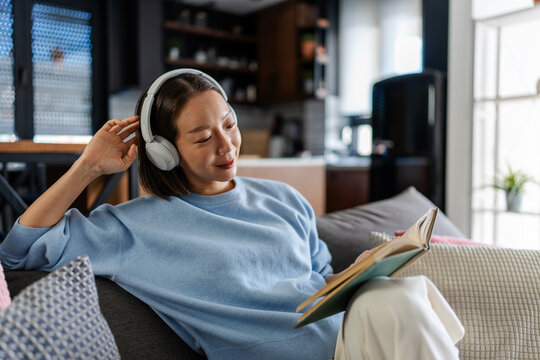 East Asian woman relaxing on a sofa at home, reading a book while wearing headphones. Peaceful, cozy moment combining music and reading for mindful relaxation and personal time.