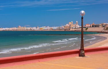 Wide coastal view of Cadiz, Spain, with sandy beach, turquoise Atlantic water, breakwaters, and the cathedral skyline under clear daylight.