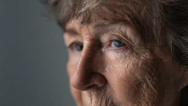 Close-up photograph of an elderly woman with short, tousled brown hair and blue eyes, showcasing wrinkles and a thoughtful expression.