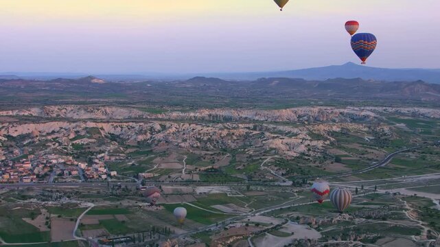 An wide shot captures numerous colorful hot air balloons as they drift across the early morning sky above the rugged, ancient valleys and distinctive "fairy chimney" rock pillars of the G&ouml;reme histori