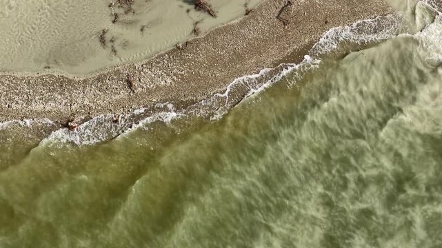 An aerial drone shot showing the moment the waves crash against the shore. An artistic blend of the water&rsquo;s texture and the natural sand and gravel formations along the shore.