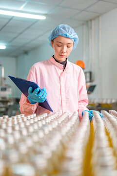 caucasian female health inspector in lab coat,hairnet and disposable gloves holding clipboard checking glass bottle of fruit juice in beverage manufacturing plant,food safety and hygiene concept