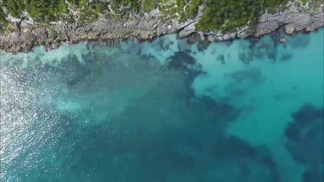 An aerial shot reveals clear turquoise waters meeting a rugged, rocky coastline with lush green vegetation, creating a beautiful and inviting seascape.