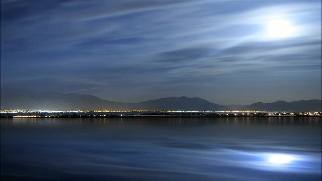 A serene body of water reflects the night sky and distant city lights, creating a peaceful scene framed by mountains under the moon's gentle glow.